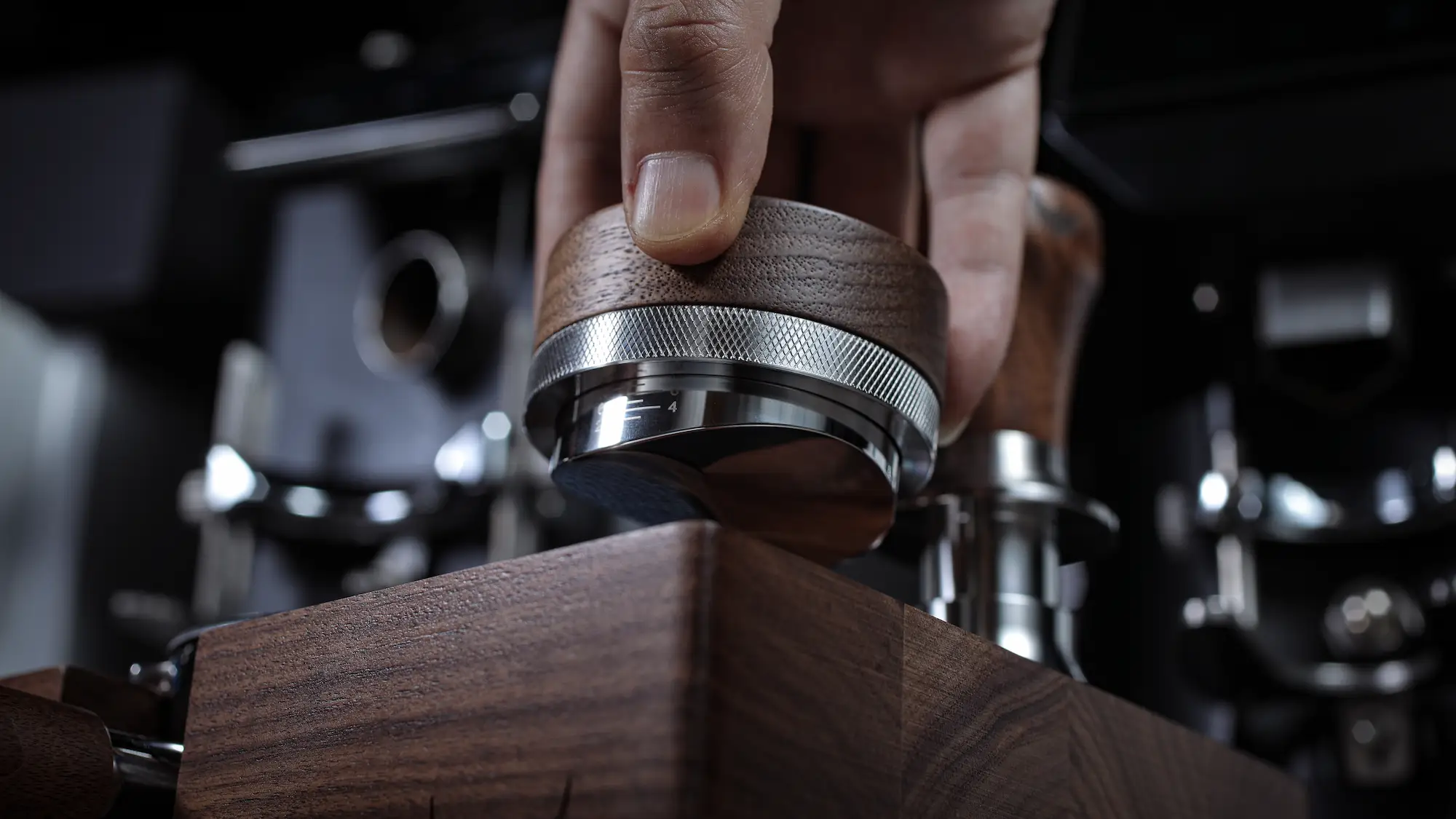 Hand holding a KNODOS espresso distributor with a walnut handle and stainless steel base over a wood tamping station