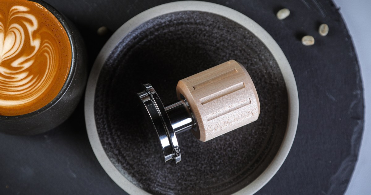 KNODOS Echo 54mm espresso tamper with a maple handle and 53.3mm stainless steel base, made for Breville 54mm portafilters, next to a latte